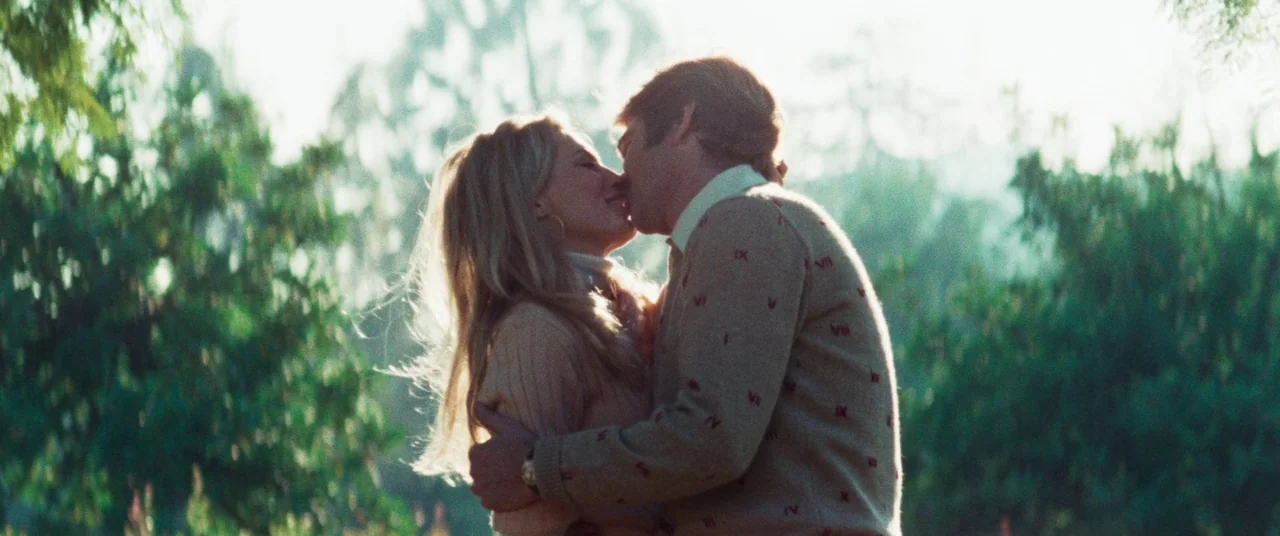 Death Game (1977) movie still: medium — A man and a woman embrace and kiss in a sunlit grassy field. Tall trees and bushes filled with green leaves…