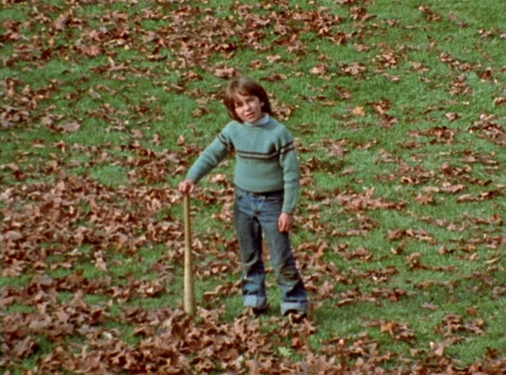Desperate Living (1977) movie still: fullbody — A green lawn is covered in scattered brown fallen leaves during the autumn season. A young boy…
