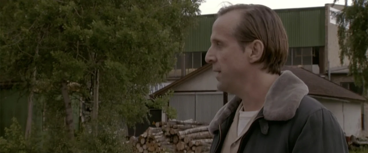 Dancer in the Dark (2000) movie still: medium — An outdoor area features stacked logs and industrial corrugated buildings in the background. A…