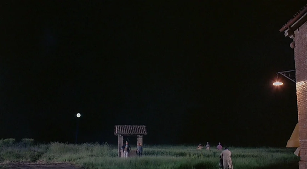 The Voice of the Moon (1990) movie still: wide — A dark night landscape shows a grassy field with a small stone-roofed well structure. Several small…