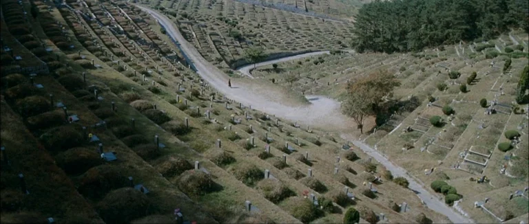 Mother (2009) movie still: establishing — A vast hillside cemetery features rows of terraced earth mounds and upright stone markers. A single small…
