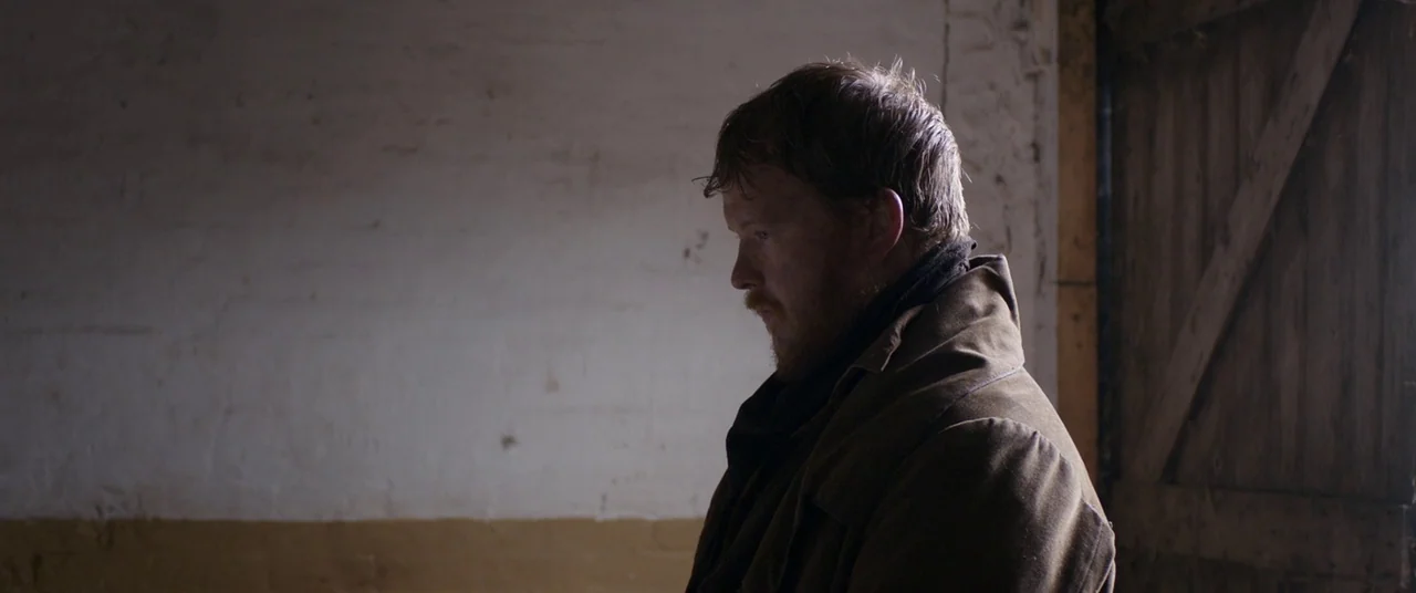 Lady Macbeth (2016) movie still: medium — The interior of a barn features a white-washed wall and wooden side door. A man stands in the soft light…