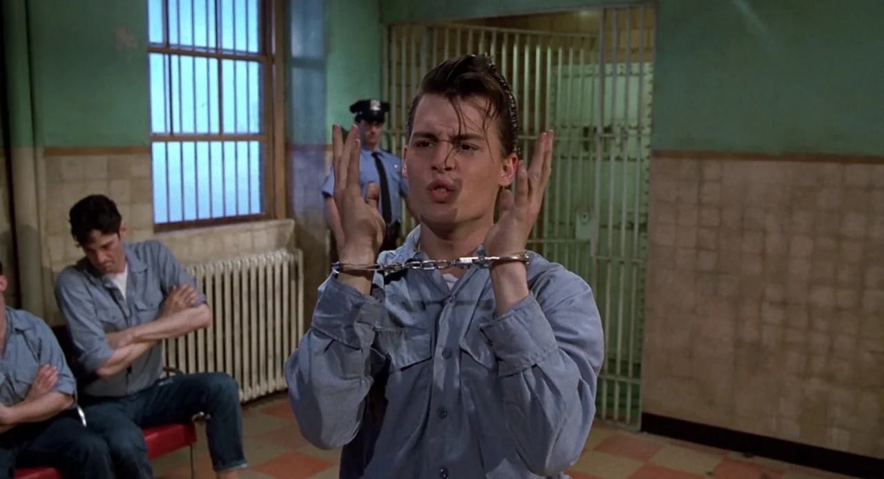 Cry Baby (1990) movie still: medium — An institutional space with pale green walls, tile flooring, and barred metal structures. A young man in a…