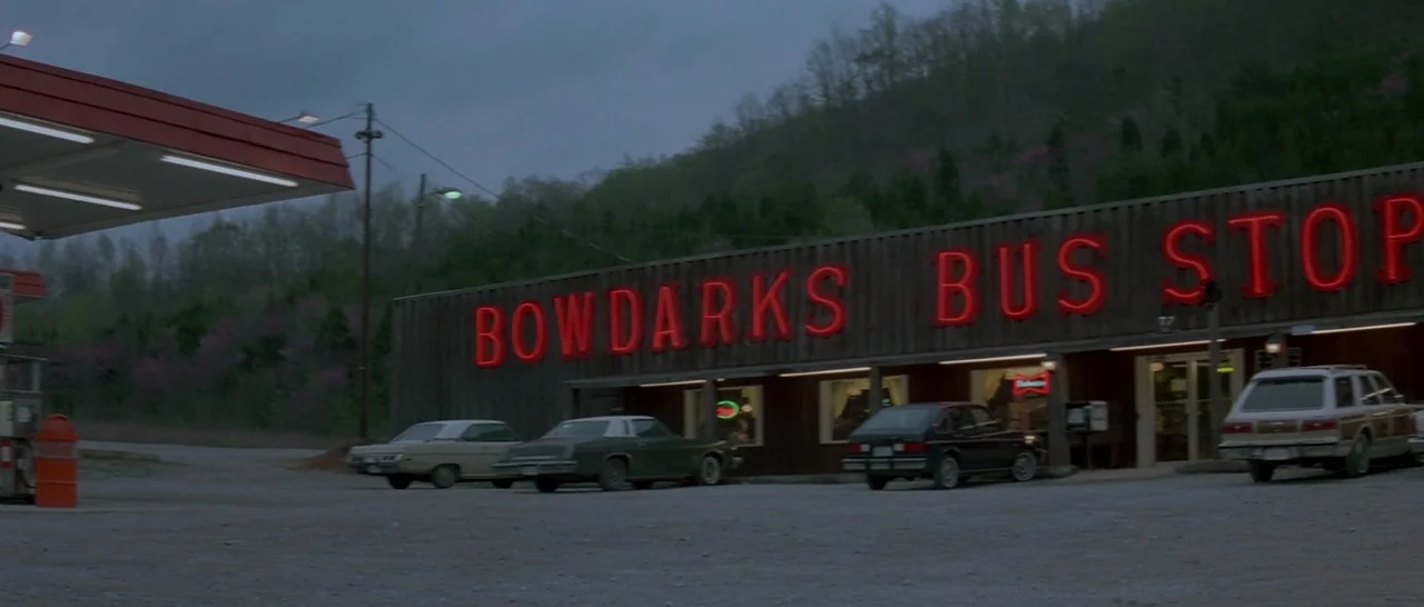 Starman (1984) movie still: wide — An isolated roadside business features a wood-paneled exterior with bright red neon signage illuminated. Several…