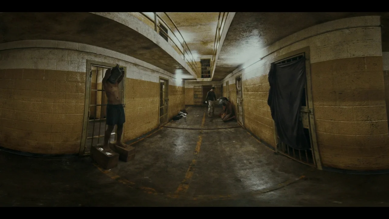The Card Counter (2021) movie still: wide — A long, dimly lit institutional corridor features yellow-painted concrete walls and gated cell doors.…
