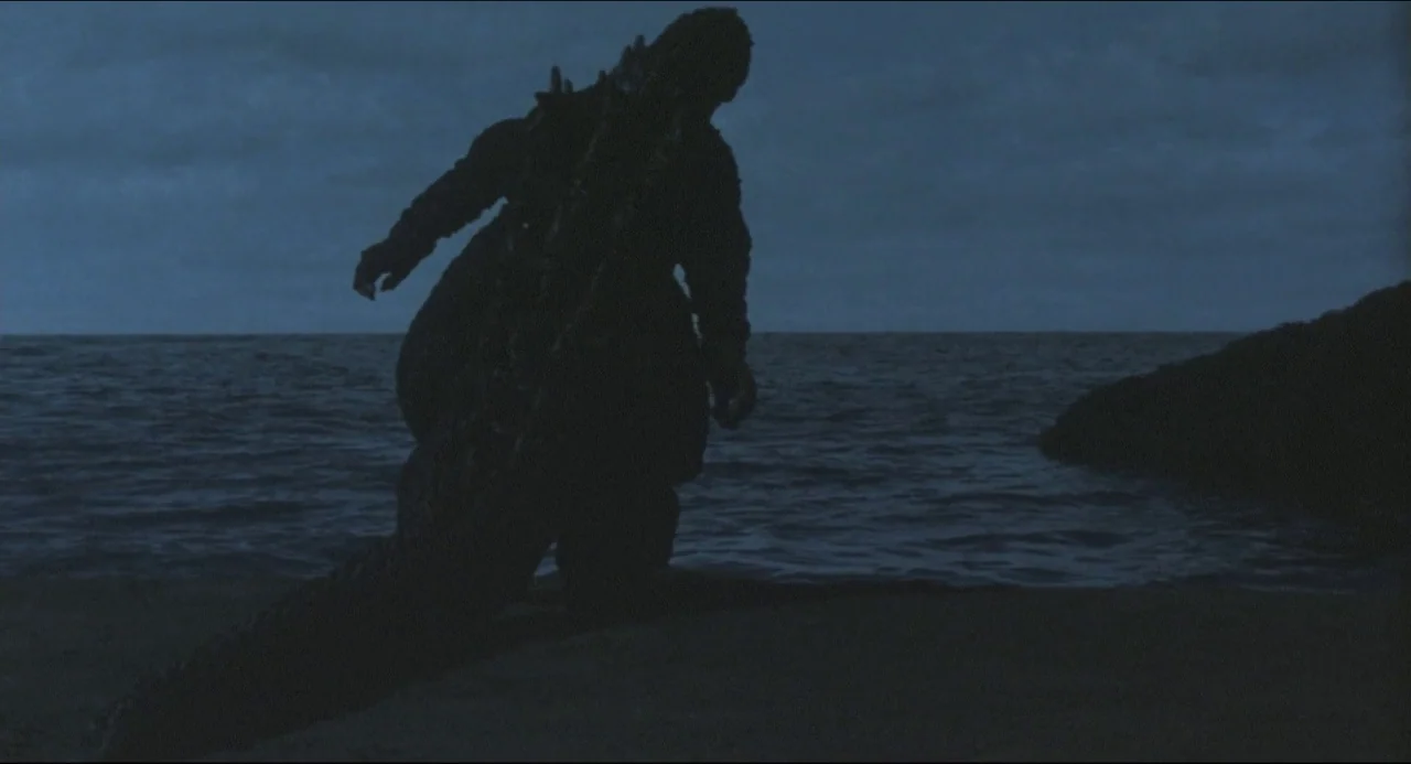 Godzilla vs. Biollante (1989) movie still: fullbody — A dark rocky shoreline at night borders a calm, deep blue sea. A large silhouette of a monster…