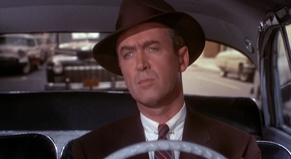 Vertigo (1958) movie still: medium — A man in a fedora drives a 1950s car through a city street. The windshield shows blurred traffic and suburban…