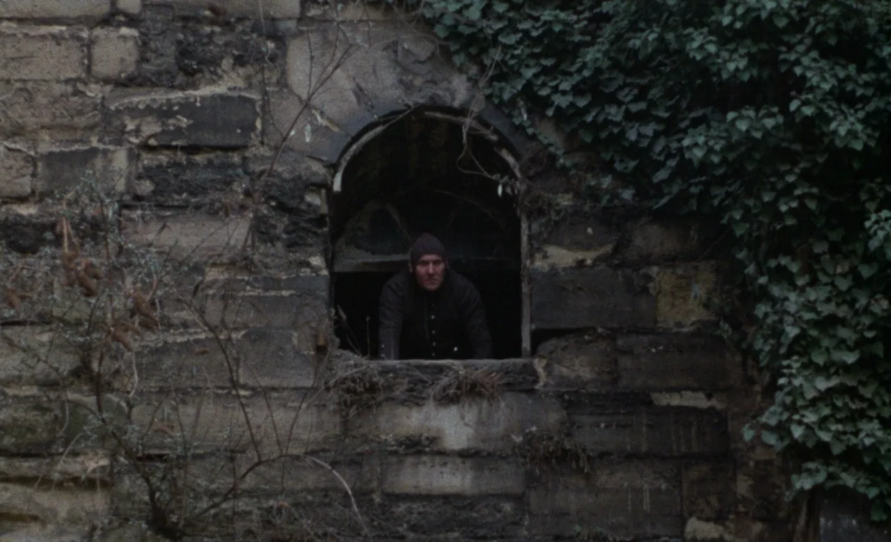 Blanche (1971) movie still: wide — An old, rough-hewn stone wall is partially covered with thick green climbing ivy. A man wearing a beanie leans…
