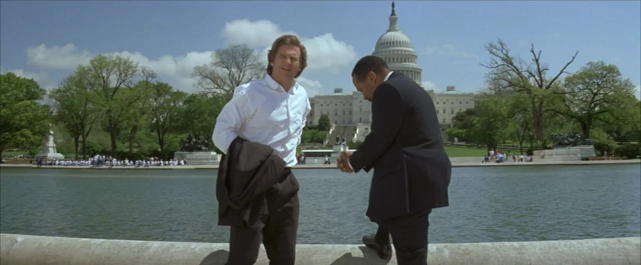 Arlington Road (1999) movie still: fullbody — A man stands with another man by a large water basin in a city park. The Capitol building is visible…