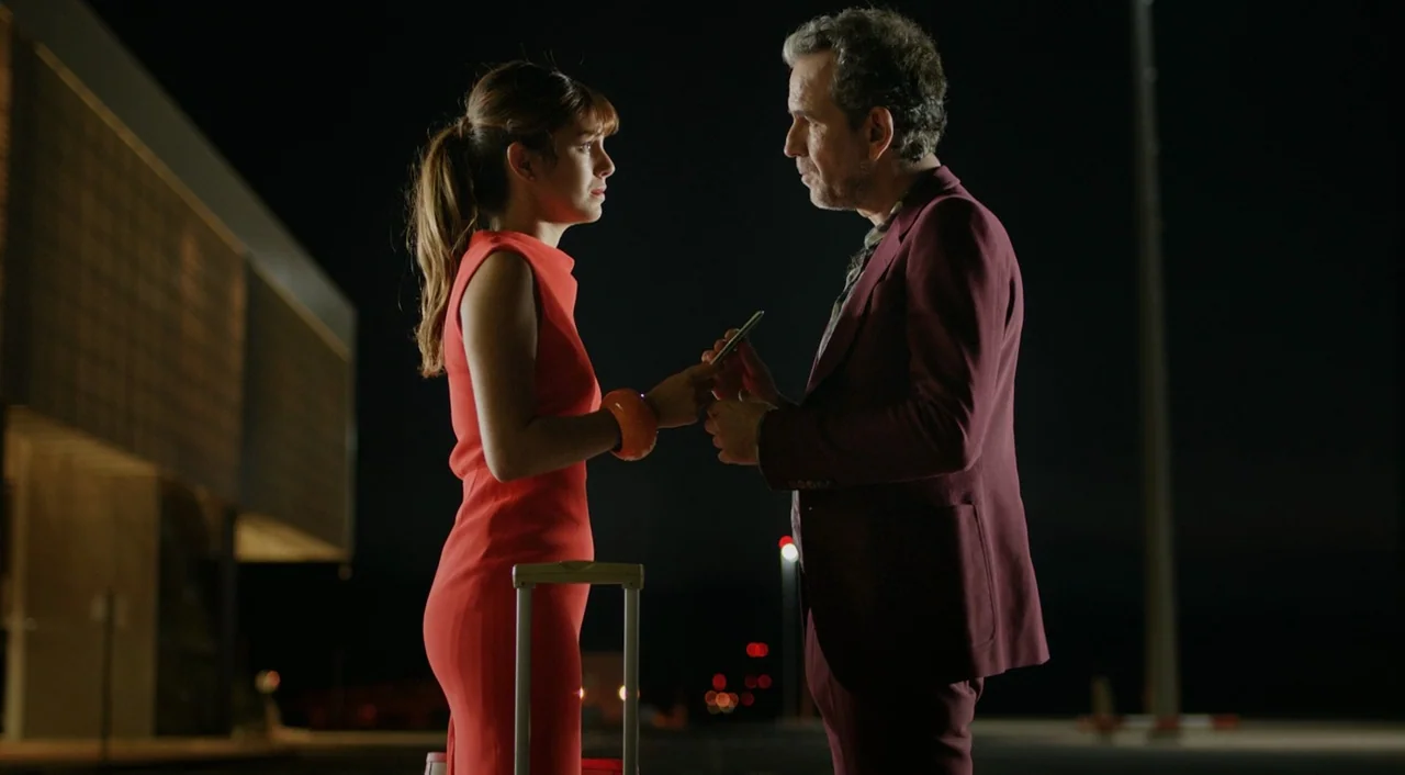 I’m So Excited! (2013) movie still: medium — An outdoor paved area at night with a modern building structure visible in the background. A woman in a…