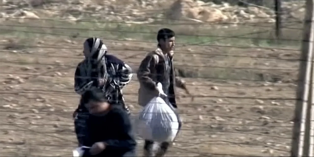 A Radical Life (2022) movie still: fullbody — A group of people navigate an arid and desolate expanse near a fence. The subjects move together…