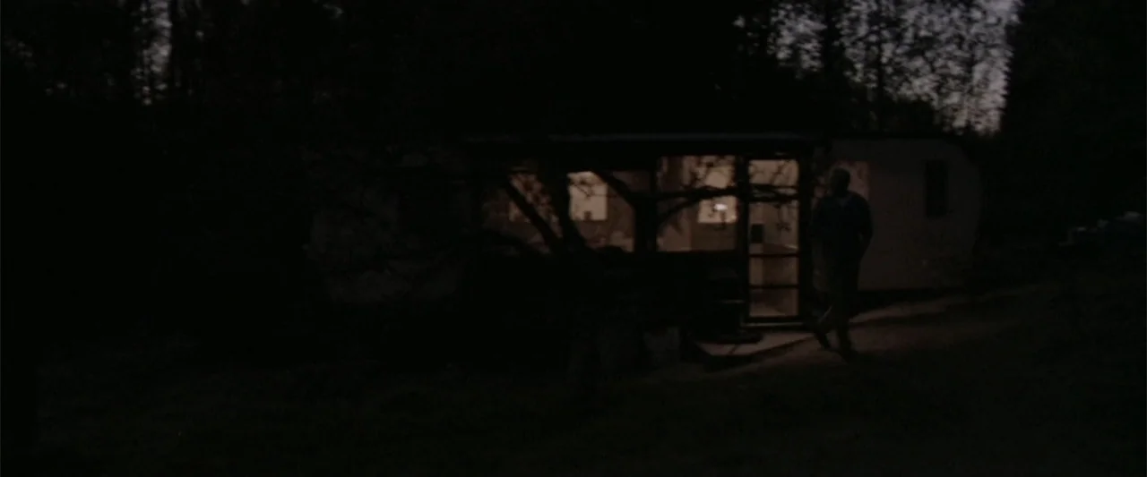 Dancer in the Dark (2000) movie still: wide — A dark, wooded area surrounds a small parked camper trailer at night. A solitary male figure walks…