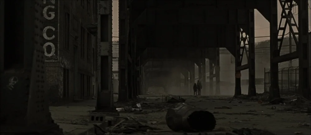 The Road (2009) movie still: wide — A desolate urban area underneath a heavy, rusted steel overpass with debris scattered throughout. Two figures…