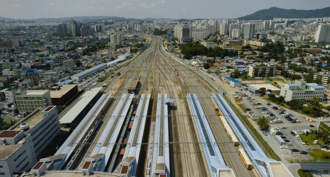 Train To Busan movie still: establishing — An aerial view of a large train station with multiple tracks and platforms. Tall apartment buildings and…