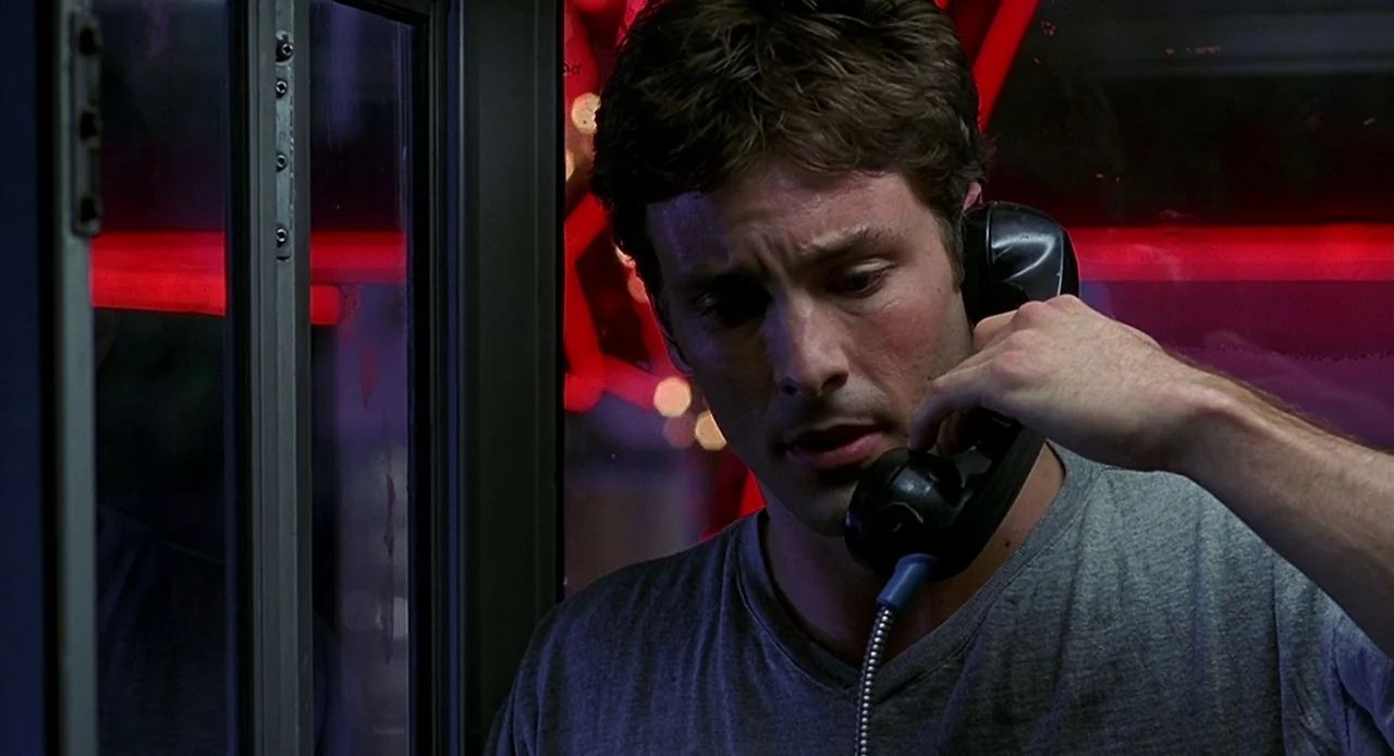 Bride of Chucky (1998) movie still: closeup — A man stands inside a glass-walled phone booth illuminated by red neon signs. He is holding a black…