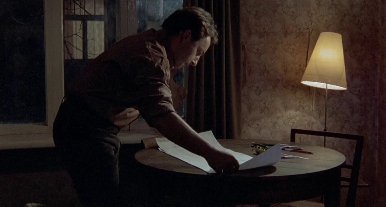 Three Colours: White (1994) movie still: medium — A dimly lit room features a dark round table against a wall with patterned floral wallpaper. A man…