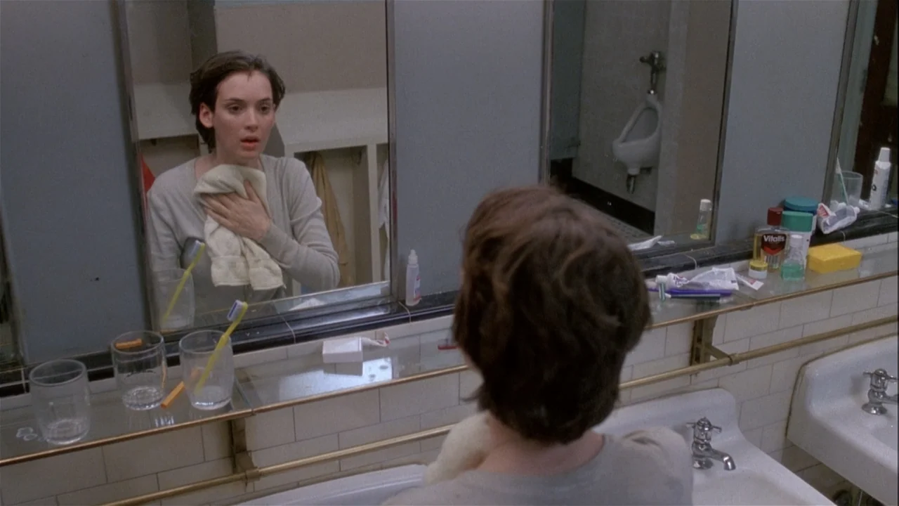 Boys (2021) movie still: medium — A public tiled bathroom features a large wall-mounted mirror above a white porcelain sink. A young woman views her…