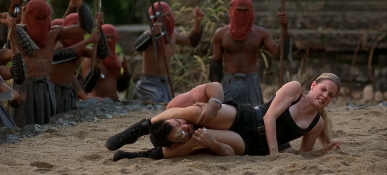Mortal Kombat (2021) movie still: fullbody — A sandy arena surrounded by several men wearing red cloth face masks and loincloths. A woman in a black…
