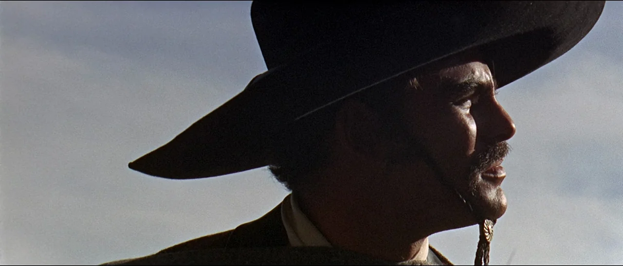 The Appaloosa (1966) movie still: closeup — An outdoor desert environment with a vast, featureless grey sky background. A man wears a black western…