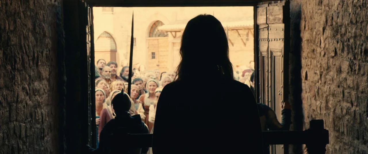 Benedetta (2021) movie still: medium — A dark stone corridor opens up to a sunlit town square filled with people. A silhouetted person stands in the…