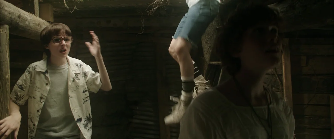 K.G.F: Chapter 2 (2022) movie still: medium — A dimly lit wooden shelter with log walls and overhead foliage. Three individuals navigate the…