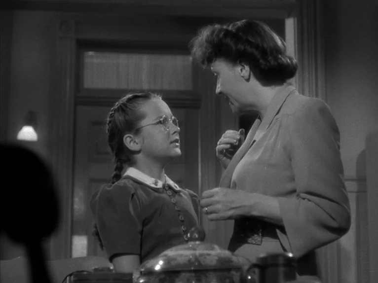 Shadow of a Doubt (1943) movie still: medium — An interior space features a woman in a suit standing before a young girl. The girl wears glasses and…