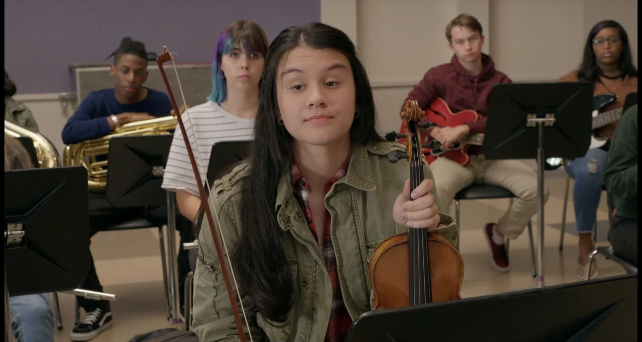 One True Loves (2023) movie still: medium — A group of students sits in a music room with instruments and music stands. A young woman in the…