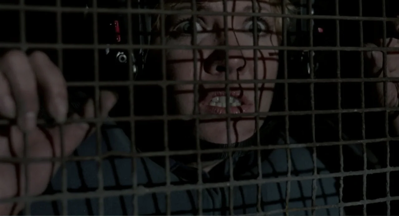 RoboCop (2014) movie still: closeup — A dark, cramped space is enclosed by a heavy iron mesh cage. A woman is positioned behind the metal grid,…