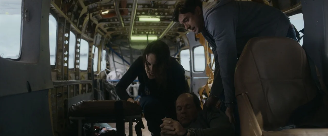 Flight Risk (2025) movie still: medium — The interior of a cramped, metallic aircraft cabin shows exposed ribs and bolted seats. A woman and a man…