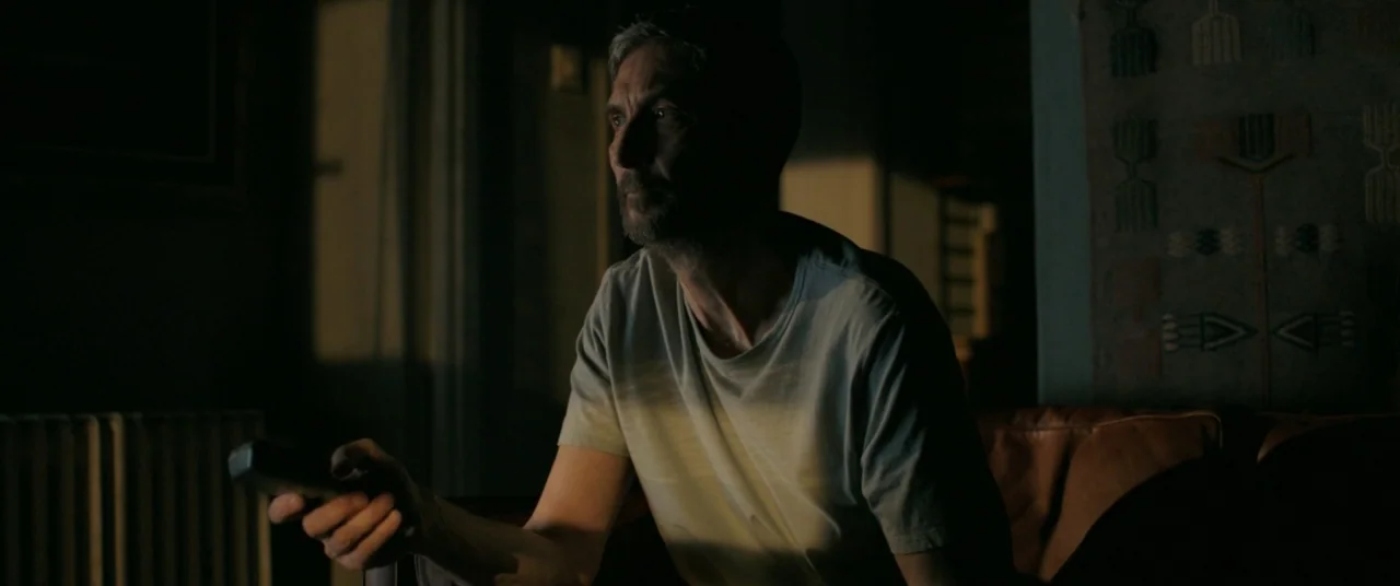 On Body and Soul (2017) movie still: medium — A dimly lit room features a man sitting on a leather sofa. The man holds a remote control while…