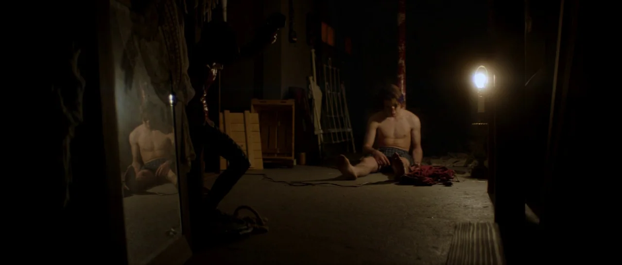 Suitable Flesh (2023) movie still: wide — A dimly lit basement features concrete flooring and scattered wooden crates and metal frames. A shirtless…