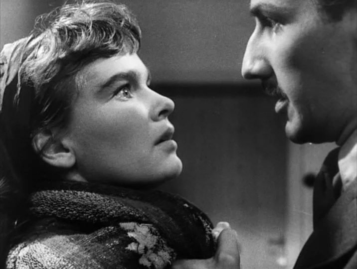 Thirst (1949) movie still: medium — A dimly lit room with blurred interior details in the background. A woman in a thick scarf looks up at a man…