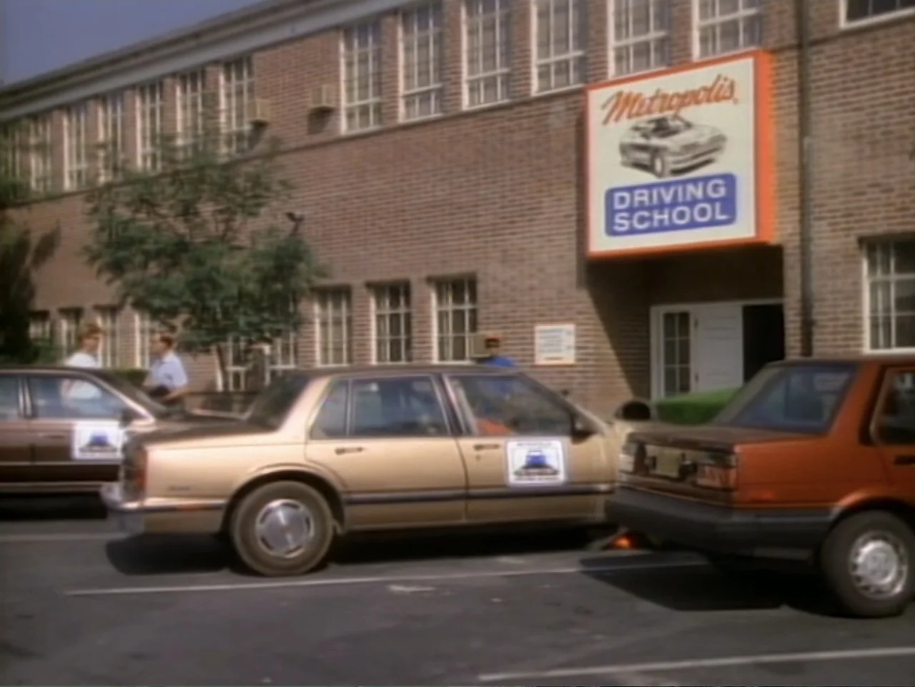 My First Love (1988) movie still: wide — A brick commercial building features a prominent sign for a driving school above the entrance. Several cars…