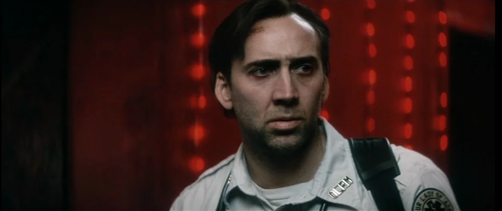 Bringing Out The Dead (1999) movie still: closeup — An interior space is bathed in darkness with out-of-focus vertical red lights behind. A man in a…