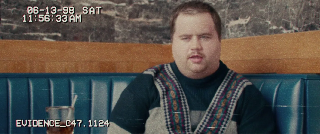 I, Tonya (2017) movie still: medium — A man sits in a blue vinyl booth inside a commercial space. He is looking forward while speaking as…