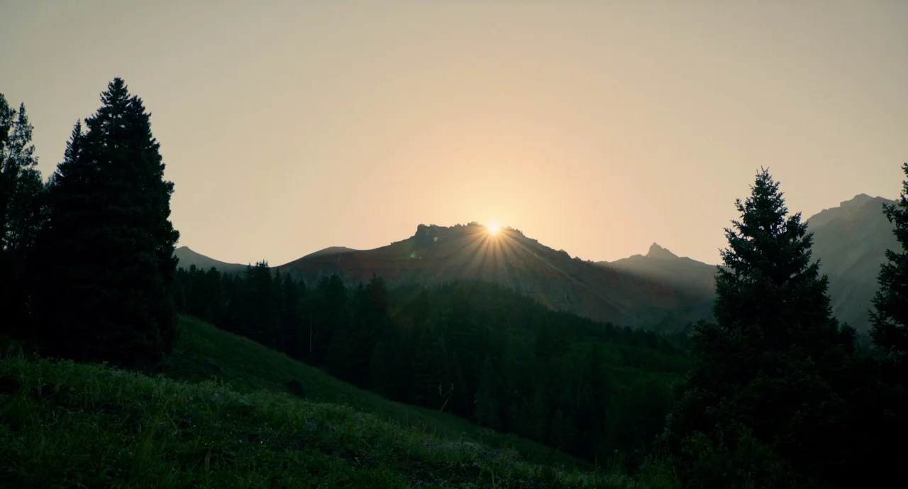 The Ballad of Buster Scruggs (2018) movie still: wide — A high mountain range at dawn features silhouettes of pine trees in the foreground. The sun…