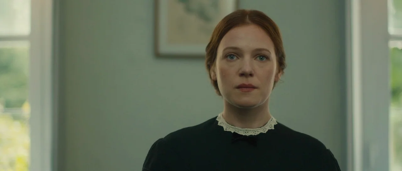A Quiet Passion (2016) movie still: closeup — A woman stands in a bright room with pale green walls and a framed print. She maintains a frontal…