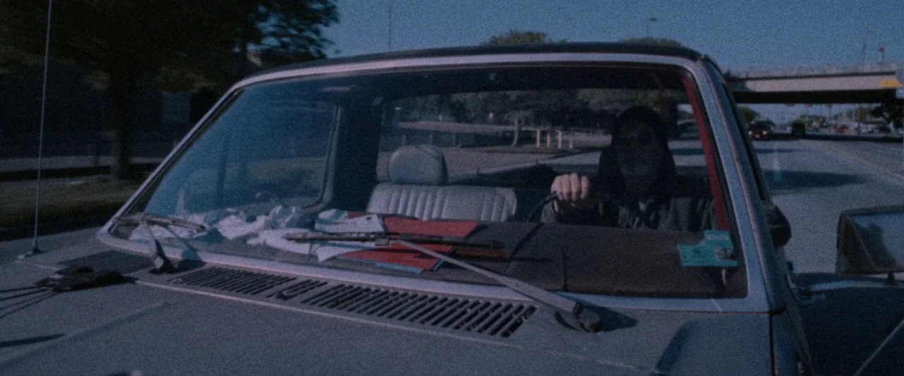 VFW movie still: medium — The interior of a moving pickup truck traveling down a sunny highway. A man sits behind the steering wheel while looking…
