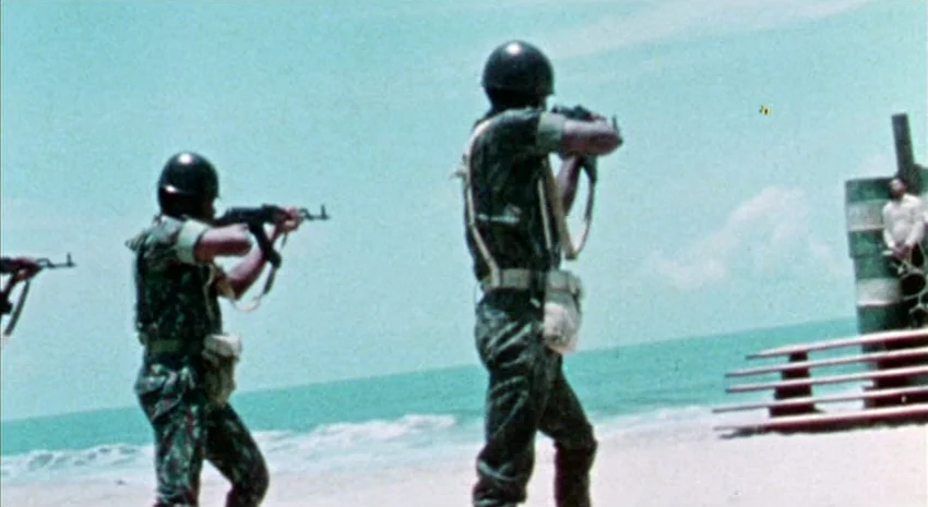 The Passenger (1975) movie still: fullbody — A bright sandy beach meets the ocean water under a clear blue sky. Two soldiers in camouflage uniforms…
