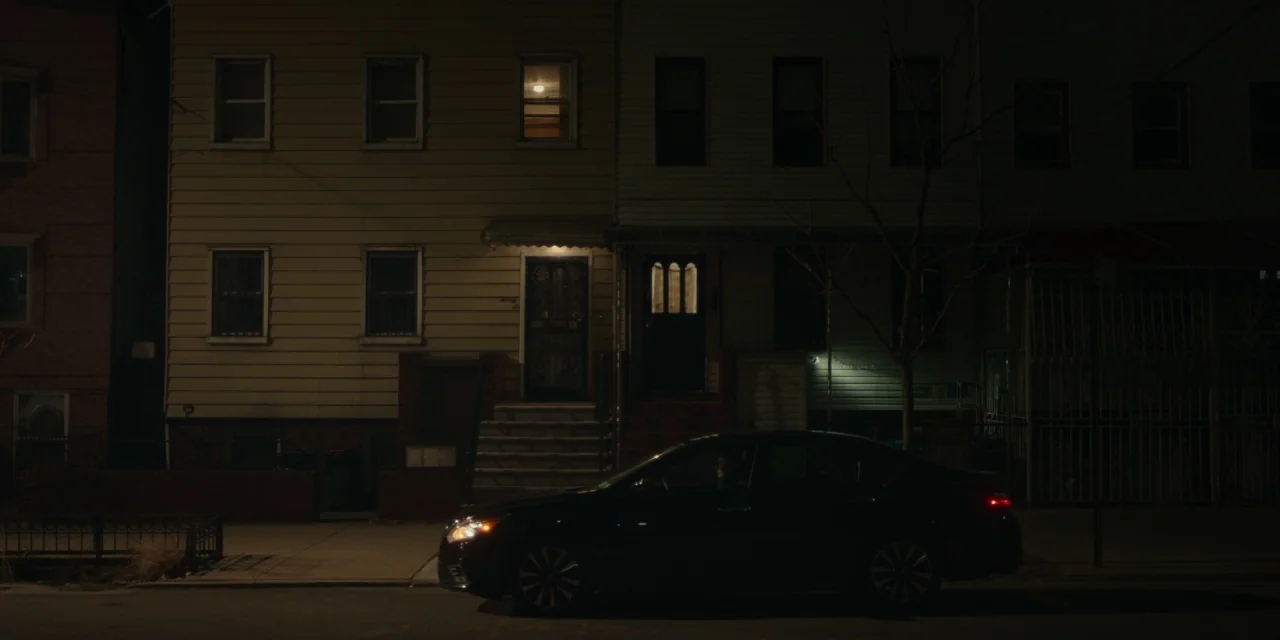 The Assistant (2019) movie still: wide — Two residential houses stand side-by-side on a quiet street at night. A black sedan is parked in front of…