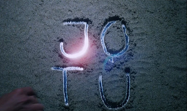 The Rose King (1986) movie still: closeup — Dark damp sand shows four hand-drawn letters glowing with an internal light source. A hand enters the…