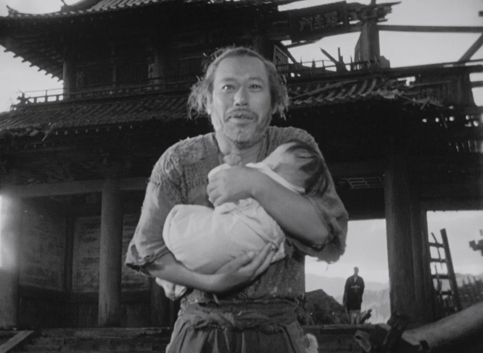 Rashomon (1950) movie still: medium — A dilapidated, traditional wooden temple gate sits in the background of a war-torn landscape. An unkempt man…