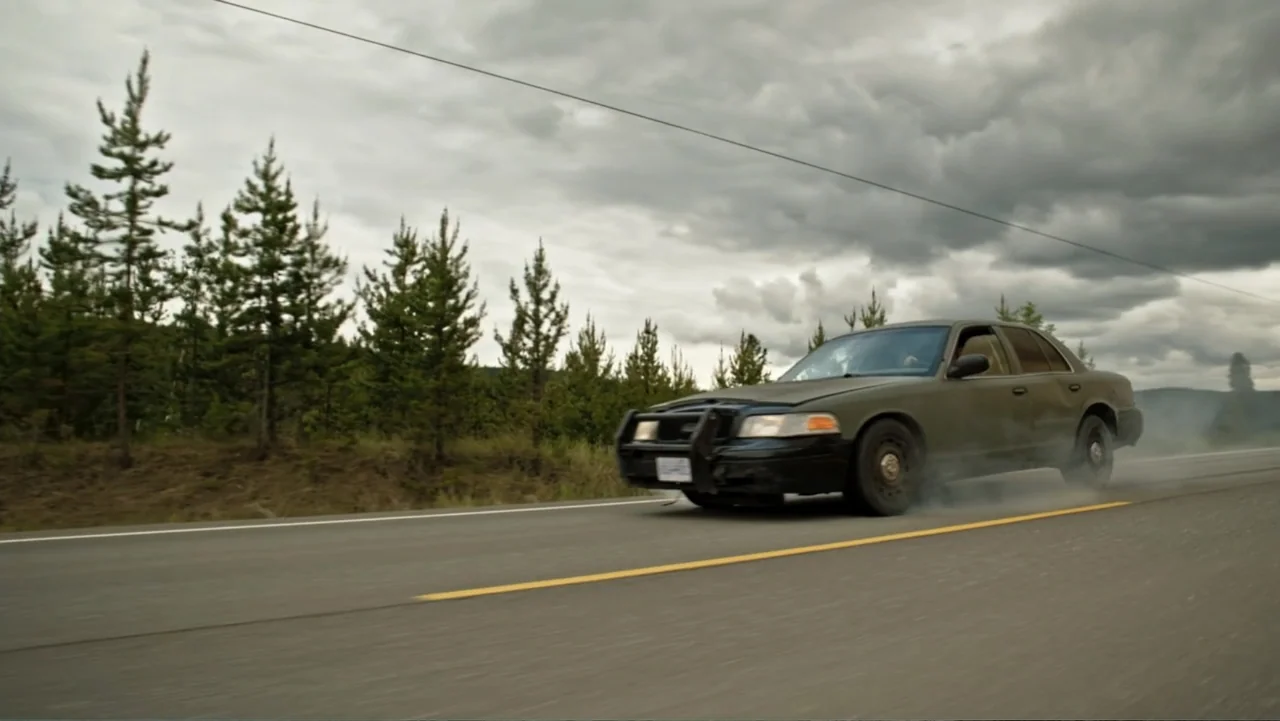 Bullet Proof (2022) movie still: medium — A dark green police interceptor sedan speeds along an asphalt road bordered by evergreen trees. The…