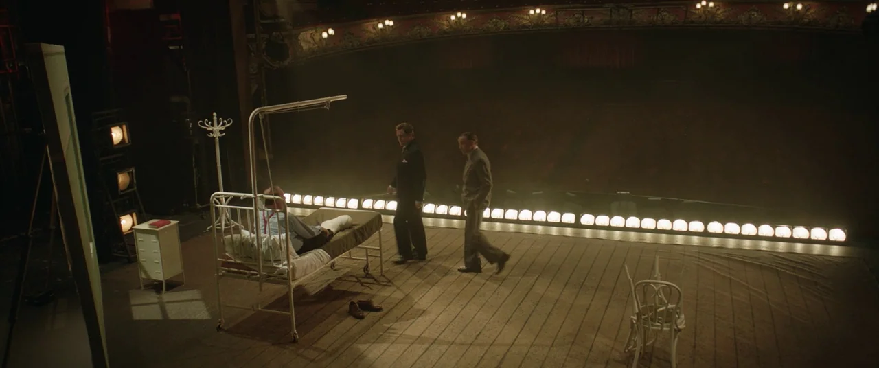 Domino (2005) movie still: wide — A bare wooden theater stage contains a lone hospital bed and scattered furniture items. Three men in mid-century…