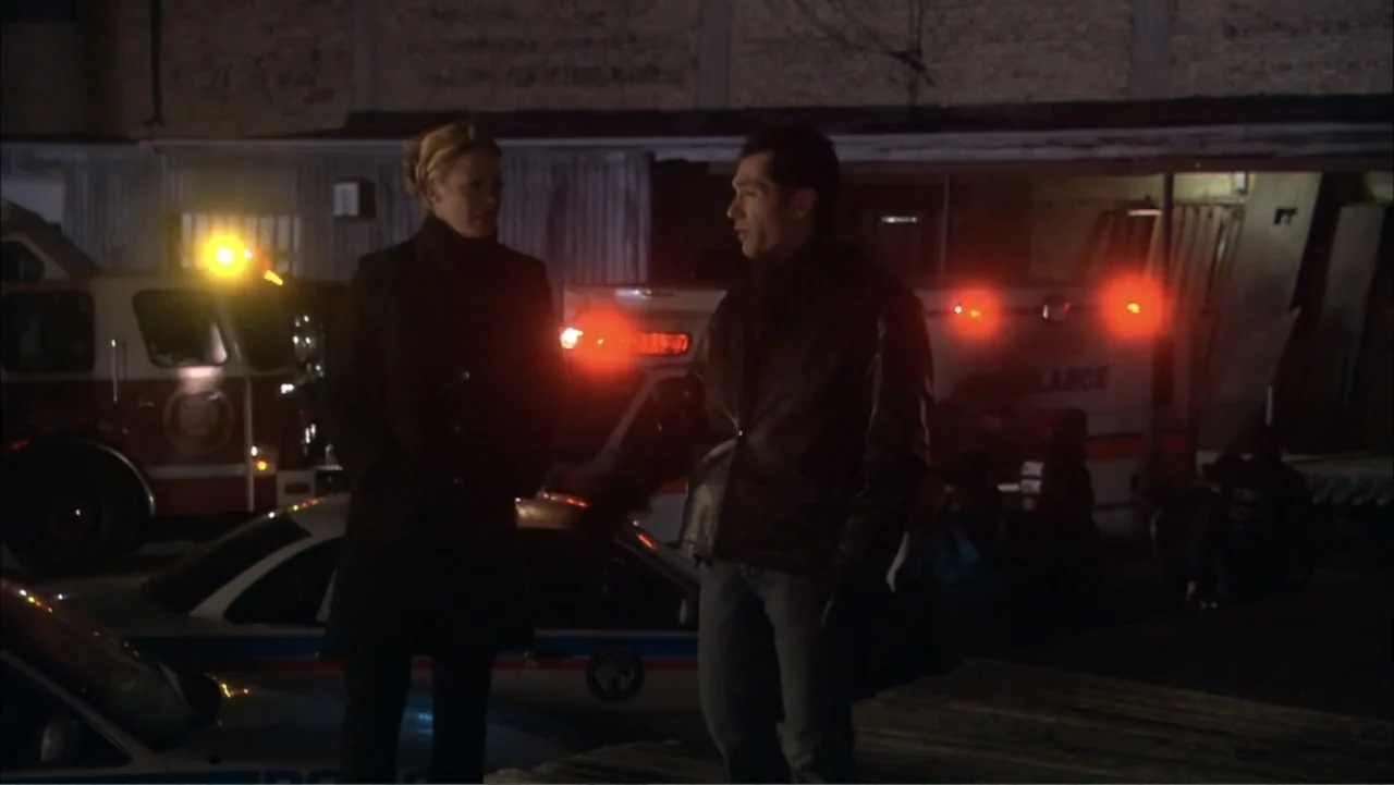 O código oculto (2025) movie still: medium — An outdoor emergency site is illuminated by bright flashing lights from parked response vehicles. A man…