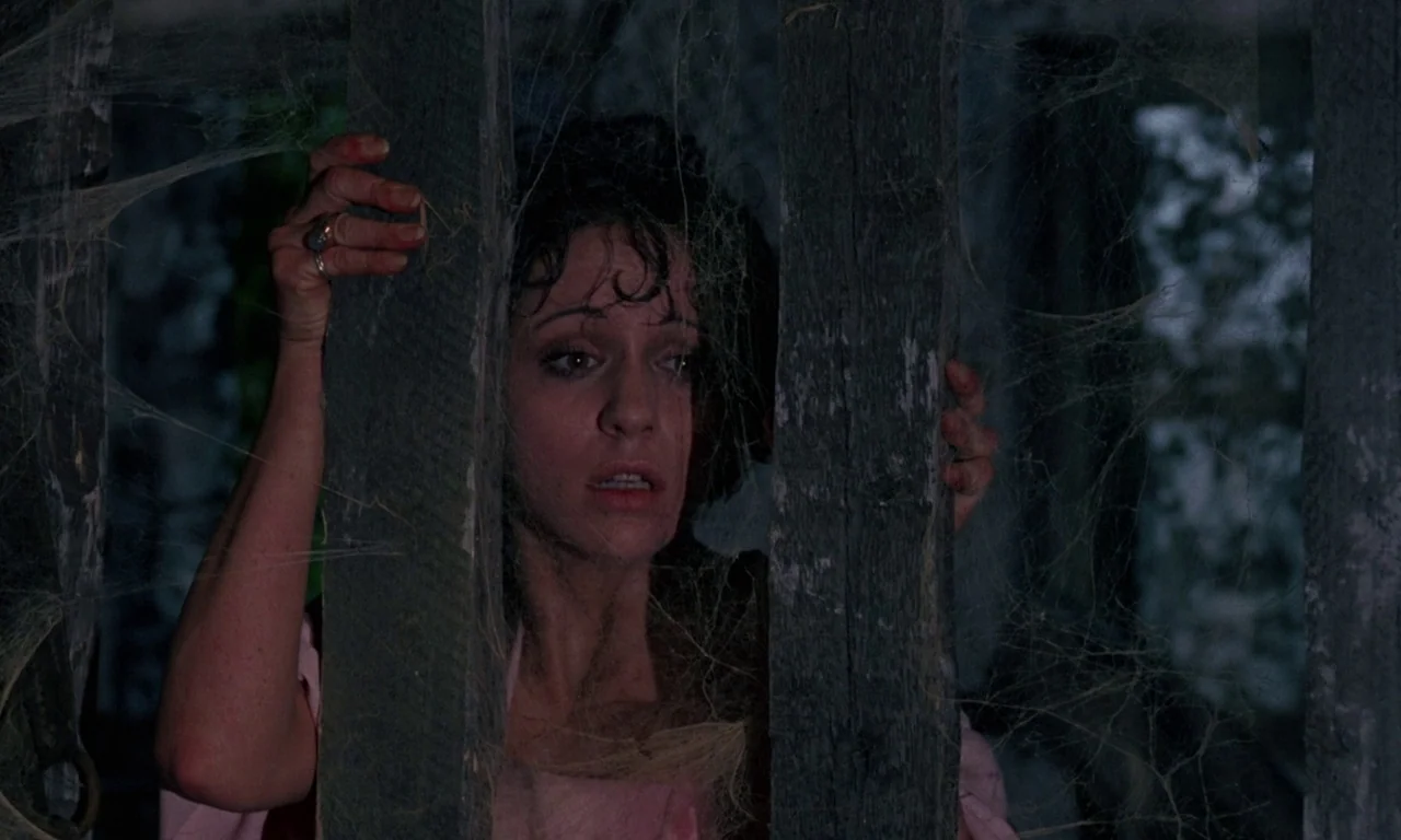 The Devil Rides Out (1968) movie still: closeup — A woman stares through vertical wooden slats covered in thick, layered cobwebs. She grips the dark…