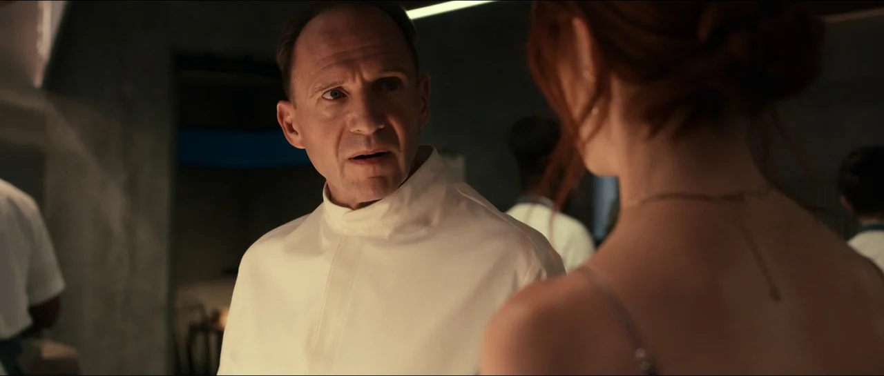 The Menu (2022) movie still: medium — A high-end professional culinary space with dark walls and subtle industrial lighting. A middle-aged man in a…