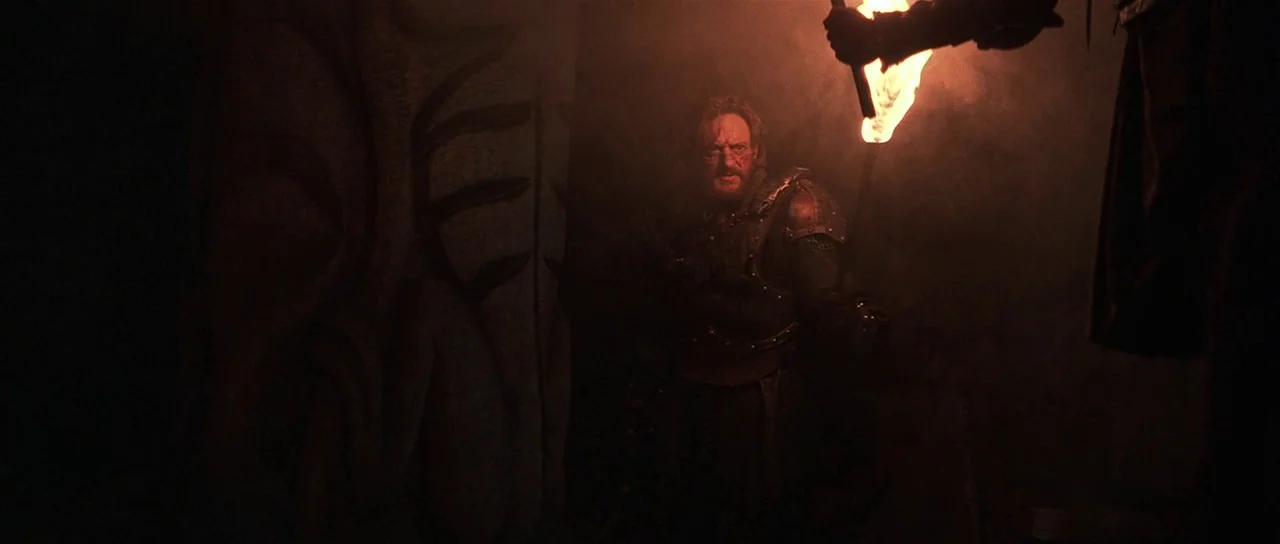The 13th Warrior (1999) movie still: medium — A dark stone room contains a large carved pillar and hazy atmosphere. A bearded man in armor holds a…