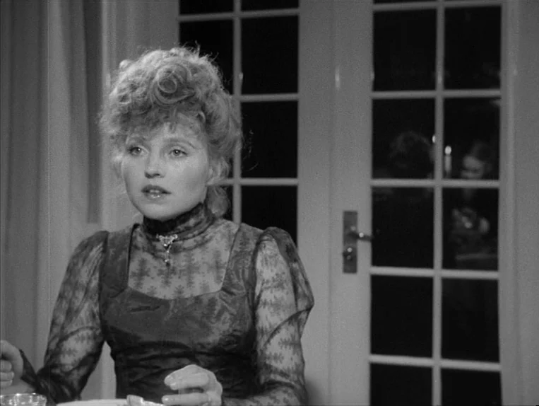 Effi Briest (1974) movie still: medium — A woman in a high-collared lace dress sits indoors at night. A white French door with glass panes is…