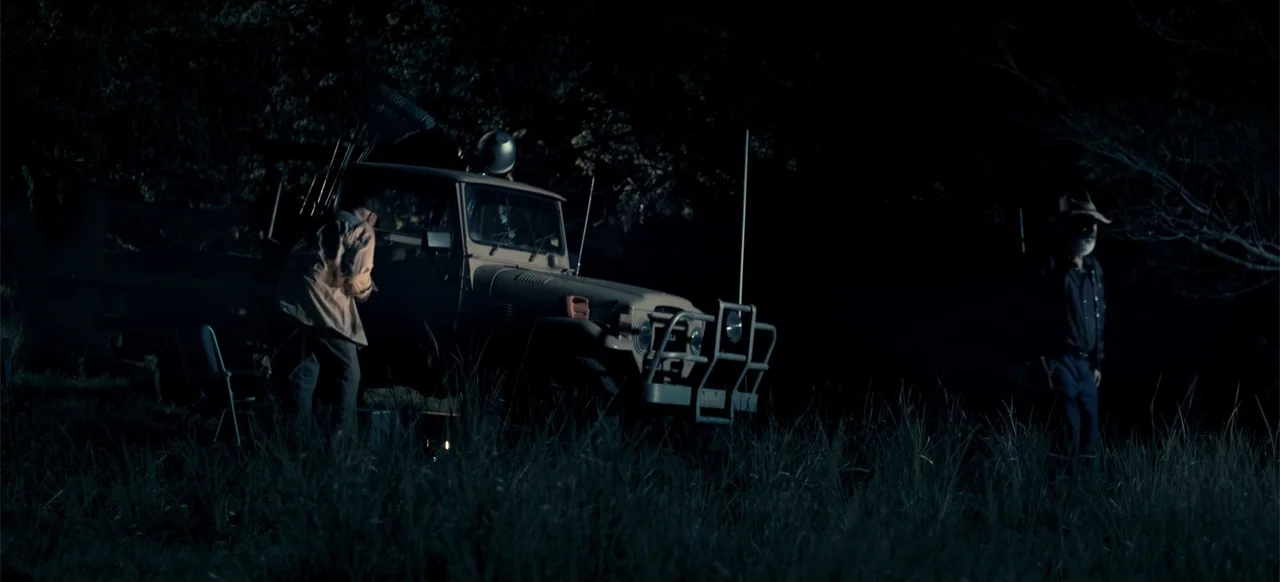 Boar (2018) movie still: wide — A dark, grassy field at night features an older truck parked in the center. Two men stand at opposite sides of the…